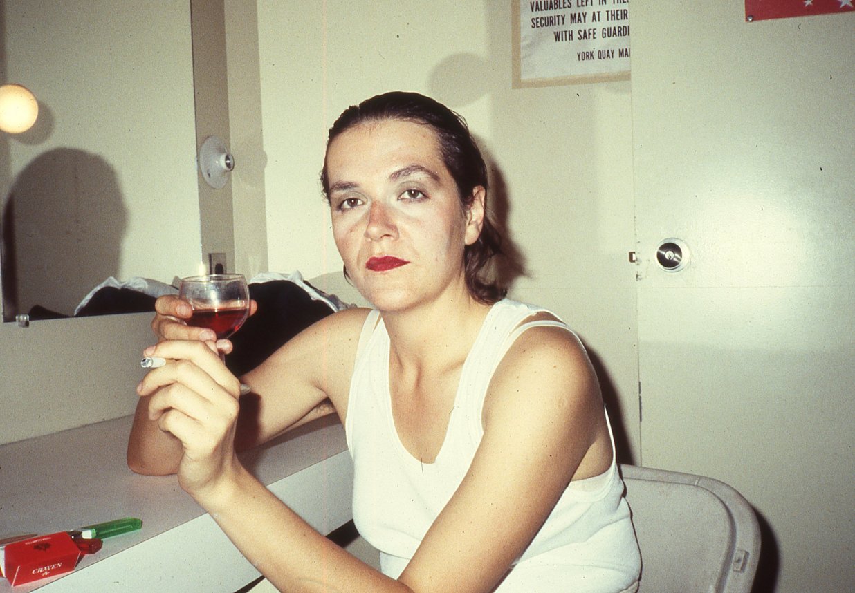 Auf dem Foto ist Margaret Dragu im Jahre 1983 in der Umkleidekabine zu sehen. Sie blickt direkt in die Kamera. In der rechten Hand h&auml;lt sie ein Glas Wein, in der linken Hand eine angez&uuml;ndete Zigarette. | © Colin Campbell
