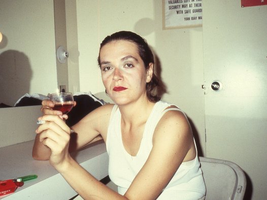 Auf dem Foto ist Margaret Dragu im Jahre 1983 in der Umkleidekabine zu sehen. Sie blickt direkt in die Kamera. In der rechten Hand hält sie ein Glas Wein, in der linken Hand eine angezündete Zigarette. | © Colin Campbell