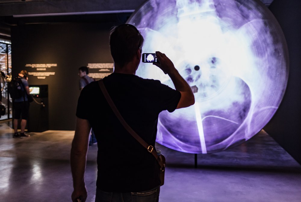 Ein Mann steht mit dem R&uuml;cken zur Kamera und macht mit seinem Smartphone ein Foto von einem Ausstellungsgegenstand, einer gro&szlig;en Schallplatte, auf welcher Visualisierungen zu sehen sind. | © X-Ray Audio Project