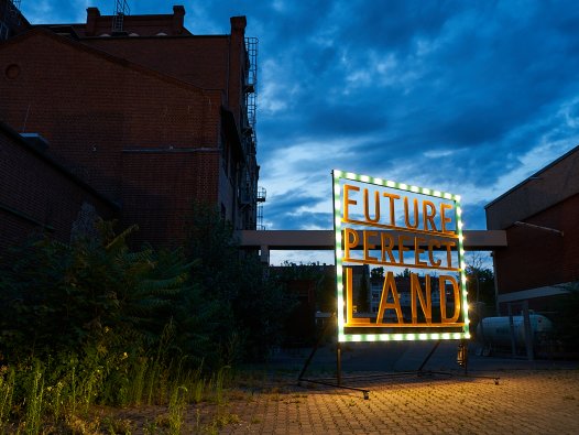 Ein Schild mit der Schrift "FuturePerfectLand" steht auf einem Industriegelände, das teilweise von Pflanzen überwachsen ist, unter einem drohenden Himmel. | © Pablo Hassmann, Magma Studio