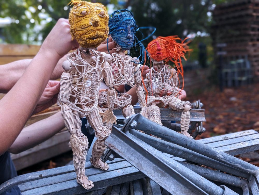 Drei Puppen aus Rattan stehen versetzt hintereinander auf einem Haufen Metallstangen im Wald. Sie tragen jeweils eine gelbe, eine blaue und eine orangene Maske. | © Artemiy Shokin